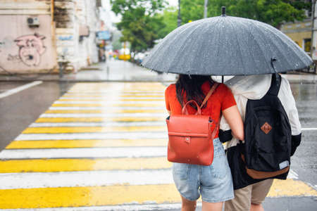 Young couple in love under one umbrella crossing the pedestrian in bad rainy weather. Rainy day. Umbrella with raindrops. City scenes in the rain.の写真素材