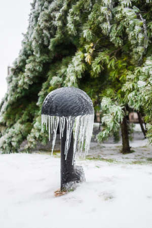 Mushroom-shaped iron lamp covered with ice and icicles. Beautiful green spruces covered with snow in the background. Ice storm, winter weather.の写真素材