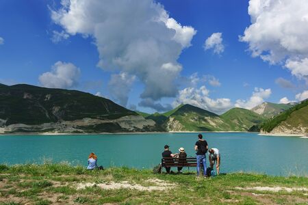 Vedensky district, Chechen Republic, Russia - June 01, 2019: Tourists sit on a bench on the shore of lake Kezenoi am. Green grass on the mountain slopes in early summerのeditorial素材