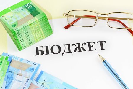 Budget-a scheme of income and expenses for a certain period of time. Money, pen, glasses and the inscription in Russian "Budget"の写真素材