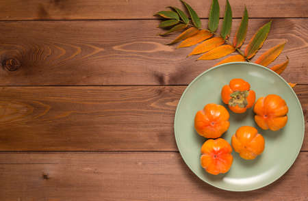 Abstract autumn still life: yellowed leaf and ripe persimmon in a plate on a brown wooden table. Flat lay. Copy space