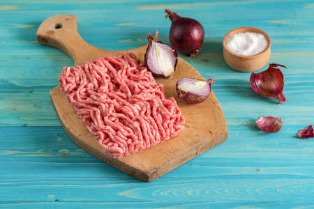Minced meat and red onions lie on a wooden cutting Board. Salt shaker on a blue wooden tableの写真素材