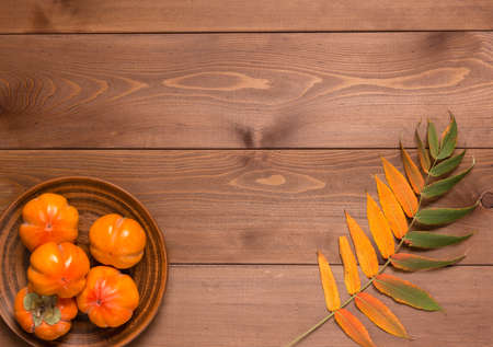 Abstract autumn still life: yellowed leaf and ripe persimmon in a clay plate on a brown wooden table. Flat lay. Copy spaceの写真素材
