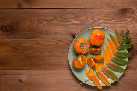 Abstract autumn still life: a beautiful yellowed leaf and ripe persimmon in a green plate on a brown...
