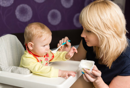 Mom feeds baby white yogurt from a jar. The child is sitting in a high chair. An apron hangs on the...