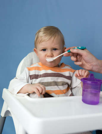A small one-year-old child with white hair is fed yogurt with a spoon. A child sits in a childs...