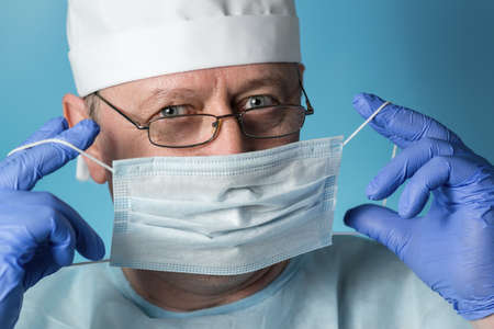 A doctor in medical clothing: a cap, gloves, dressing gown and glasses puts on or removes a protective mask. Close-up, look at the cameraの写真素材