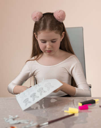 A school-age girl is sitting at a table and is concentrating on cutting jewelry and gifts out of...