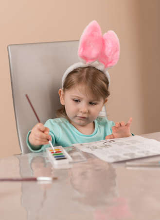 A little girl in a cute headband with pink bunny ears draws with a brush with watercolor paintsの写真素材