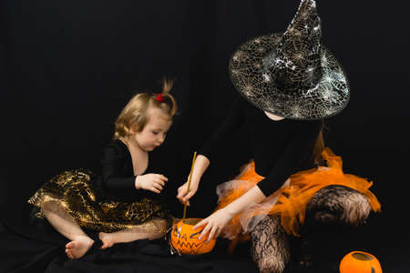 Two sisters in witch costumes, cooking a magic potion in a bucket-pumpkin. A black blouse, a hat with cobwebs, an orange skirt. Black backgroundの写真素材