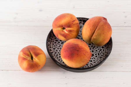 Four fresh ripe juicy peaches are lying in a black clay plate on a white wooden tableの写真素材
