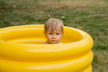 A serious little girl with a wet head in a yellow inflatable pool on a green lawn background. Toddlerの写真素材