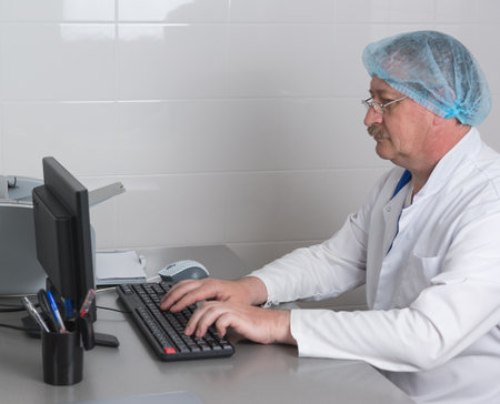 An elderly male doctor in glasses and medical clothes is sitting at his desk in office and typing examination report on computerの写真素材