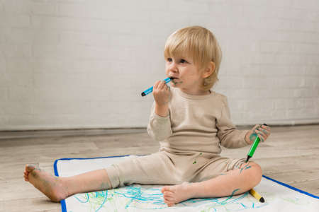 A little cheerful girl in pajamas is sitting on the floor and drawing with water markers on a...