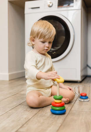 A small child sits on the floor of the house and collects a wooden toy on a stick, a pyramid of colorful rings. Lifestyleの写真素材