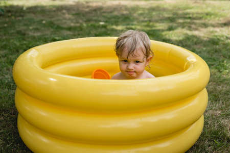 A cheerful girl with a toy and wet hair in a yellow inflatable pool on a green lawn. A glance at the camera. Toddlerの写真素材