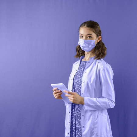 A beautiful student girl in a medical suit and mask with a notebook in her hands stands on a purple...