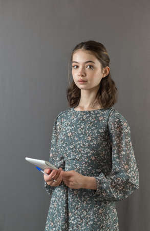 A beautiful serious successful girl in a dress stands against a gray wall with a notebook and pen in her handsの写真素材