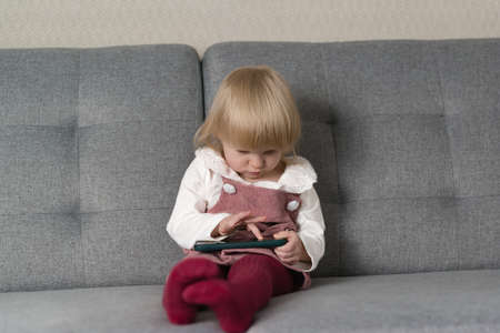 A little beautiful girl is sitting at home on the couch and playing with her smartphoneの写真素材