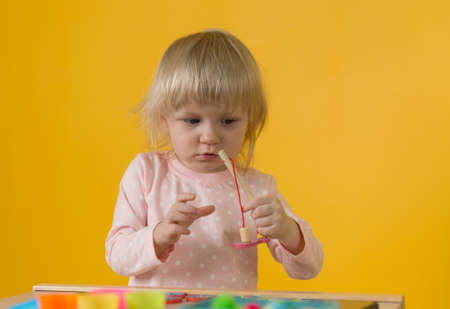 A girl in pink pajamas against a yellow wall enthusiastically plays a magnetic fishing toy with a wooden fishing rod. Development of motor skills, attention, coordination, perception of shapes, colorsの写真素材
