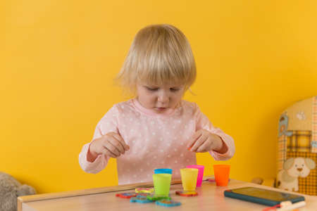 A girl in pink pajamas at a table against a yellow wall is playing sorting balls into cups. Development of motor skills, attention, coordination, perception of shapes and colorsの写真素材
