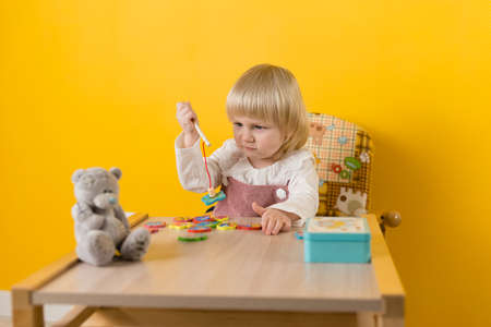 A girl in a dress is sitting at a table against a yellow wall and enthusiastically playing fishing with a magnetic toy. Development of motor skills, attention, coordination, perception of shapes, colorsの写真素材