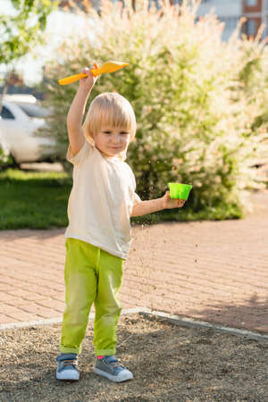 A blond, cheerful child stands alone on a playground in the city and plays with sand and toys. Sunny summerの写真素材
