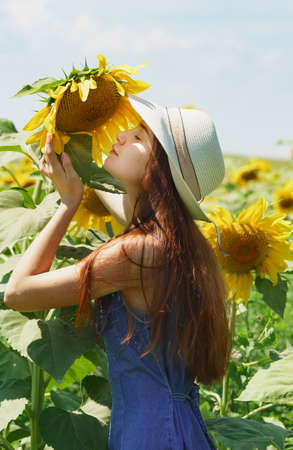 A girl in a summer hat sniffs a flower of a tall sunflower. Sultry summer in the fieldの写真素材