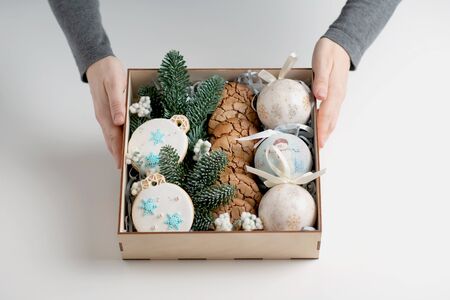 Woman's hands are holding a Christmas wooden box with cookies inside with gingerbread in shape of ball, with branch of fir on white table.の写真素材