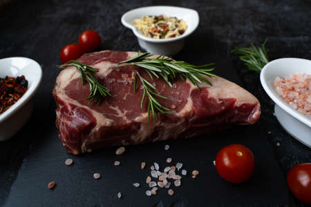 Raw rib eye steak beef with rosemary and tomato on a slate square board on a wooden background.の写真素材