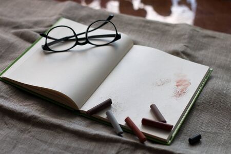 black glasses on the clean white pages of notebook for sketch paint and drawings with pastel on it on the flax background on the table near the windowの写真素材