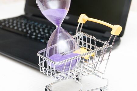 hourglass in a shopping trolley next to a laptop on a gray table, online shopping concept