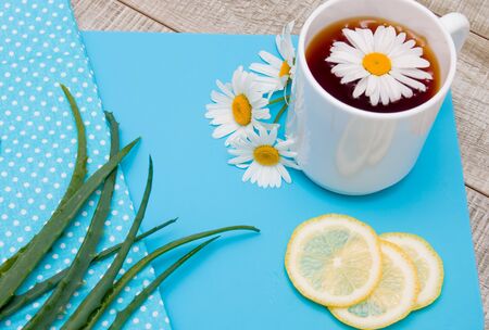 natural medicine, white cup with chamomile tea on a blue cutting board with daisy flowers, chopped lemon, aloe and polka dot fabric on a gray wooden tableの写真素材