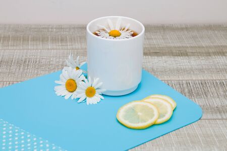 white cup with chamomile tea on a blue cutting board with chamomile flowers, sliced lemon and polka-dot cloth on a gray wooden tableの写真素材