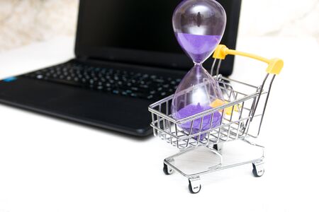 hourglass in a shopping trolley next to a laptop on a gray table, online shopping concept