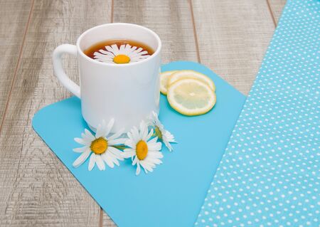 white cup with chamomile tea on a blue cutting board with chamomile flowers, sliced lemon and polka-dot cloth on a gray wooden table copy spaceの写真素材