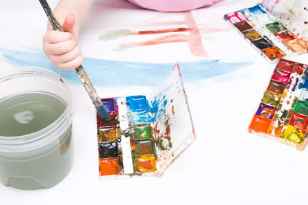the child paints with a brush watercolors on a white sheet, a table, a copy space, several packages of watercolor, a large art brush in his hand, a can of waterの写真素材
