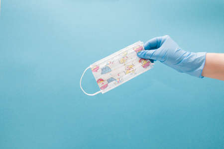 female hand holds children's medical protective mask with drawings on a blue background copy space,...