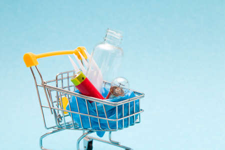 disposable plastic white plug, battery, light bulb and bottle in a shopping trolley on a blue background copy space, we buy trash, zero waste lifestyle conceptの写真素材