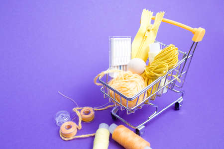 shopping trolley with various accessories and accessories for sewing, yellow threads of different colors, needles for a sewing machine, a ball of thread on a purple background copy space, hobby shopping conceptの写真素材
