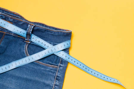 blue measuring tape is threaded into jeans instead of a belt, blue jeans on a yellow background,...