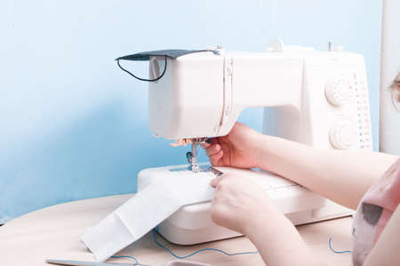 girl sews on a sewing machine a protective mask for face from cotton gray fabric, blue background,...