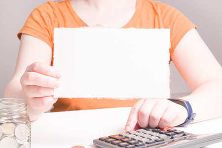 woman holds empty white cardboard, copy space, coins, bank cards on the table and in a glass jar,...