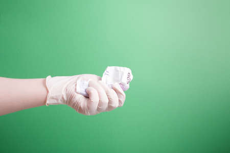 female hand in a rubber white medical disposable glove holds a crumpled 500 euro bill green background copy spaceの写真素材