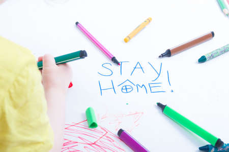 a small child in a yellow T-shirt draws on paper what to do with the child during quarantine, the development of preschool children, the inscription stay home on a piece of paper, focus on the inscriptionの写真素材