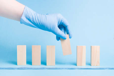 hand in a rubber disposable blue medical glove takes a domino block, dominoes are lined up, blue background, copy space, social distance concept, isolation of a sick person from societyの写真素材