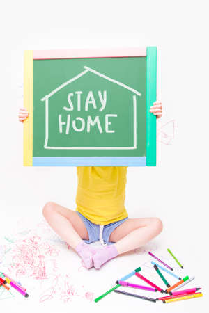 a child in a yellow T-shirt and blue shorts holds a green chalkboard with the words stay home, white background painted with children's drawings, quarantine activities with childrenの写真素材