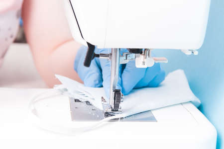 a woman in blue disposable medical gloves sews on a sewing machine a protective face mask made of cotton gray fabric, blue background, the process of sewing masks, focus on the sewing machineの写真素材