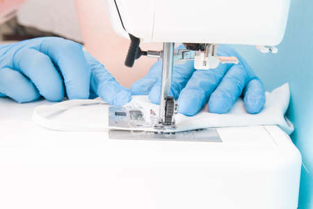 a woman in blue disposable medical gloves sews on a sewing machine a protective face mask made of cotton gray fabric, blue background, the process of sewing masks, focus on the sewing machineの写真素材