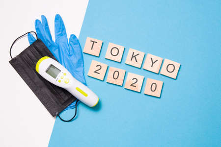 blue disposable medical glove, black face mask, infrared thermometer for non-contact temperature measurement and wooden squares with the inscription Tokyo 2020, blue backgroundの写真素材